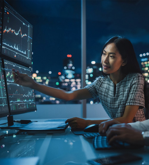  Individual pointing at financial charts displayed on multiple monitors in an office at night.