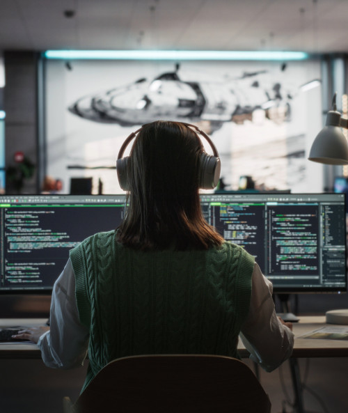  Individual wearing headphones, seated at a desk with multiple monitors showing code.