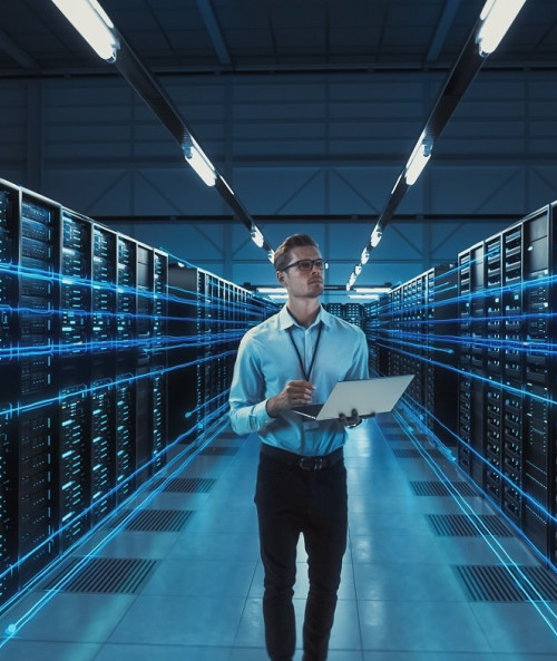 Person walking in a server room with a laptop: Individual walking through a server room, holding a laptop, surrounded by rows of servers.